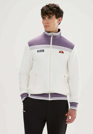 Junger Mann trägt eine weiße und lila Sportjacke mit Reißverschluss und schwarze Hose, steht mit einer Hand in der Tasche vor einem schlichten Hintergrund.