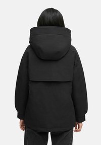 Zwarte pufferjas met een grote kap, vierkante silhouet en ribgebreide manchetten. Heeft een zachte structuur en een horizontale klepspecifiek detail op de rug.