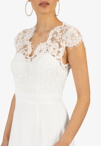 Weißes Kleid mit einem Spitzenoberteil mit Blumenmustern, kurzen Flügelärmeln und einem glatten, fließenden Rock. Strukturelle Details entlang des Ausschnitts.
