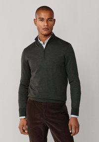 Dunkelgrüner Reißverschluss-Pullover mit gerippten Bündchen und Saum, über einem hellblauen Hemd mit Kragen getragen, kombiniert mit dunkelbraunen Hosen.