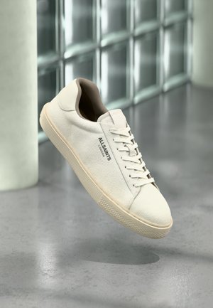 Zapatilla de lona blanca con suela y cordones beige, logo "AllSaints London" a un lado, flotando sobre un suelo gris pulido cerca de bloques de cristal.