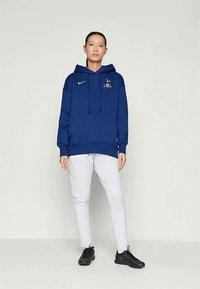 Blå bomullshoodie med snøring, frontlomme, Nike-logo i gult og brodert grafikk/design på brystet, sammen med hvite bukser.