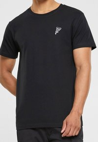 Černé bavlněné tričko s malou bílou grafikou pizzy na levé části hrudi. Klasický střih s kulatým výstřihem a krátkými rukávy.