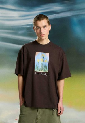 Młody mężczyzna z krótkimi włosami nosi ciemną, dużą koszulkę z nadrukiem żółtego kwiatu opatrzonego podpisem "Claude Monet", stojąc na tle rozmytego, wielokolorowego tła.