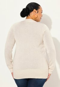 Beige långärmad tröja med ribbad nederkant och ärmslut. Lättviktig stickad textur, medellång design, bäruv över mörkblå jeans.