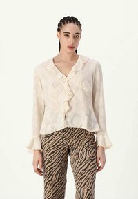 Blusa de creme claro com gola plissada e fecho de botões, apresentando mangas longas com punhos plissados. Combinada com calças listradas em castanho e preto.