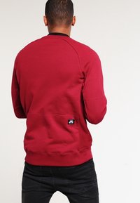 Bordeauxtröja i bomullsblandning, med rund halsringning, ribbade ärmmuddar och en liten logotypapplikation på den nedre delen av ryggen.