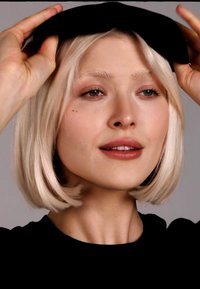 Mujer con cabello rubio corto ajustándose una boina negra, vistiendo una camiseta negra, con un fondo gris neutro.