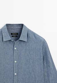 Chemise en lin bleu clair à boutons avec col classique, boutons blancs et une étiquette Massimo Dutti visible à l’intérieur du col.