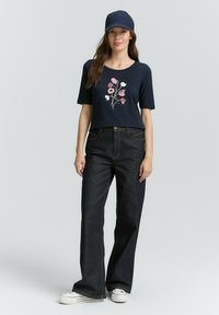 Marineblaues T-Shirt mit kurzen Ärmeln und floralem Stickerei, kombiniert mit hochgeschnittenen dunklen Denim-Hosen und weißen Sneakers. Eine schlichte Mütze rundet den Look ab.