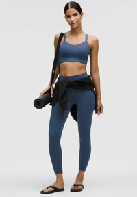 Mujer vistiendo un sujetador deportivo azul y leggings, sosteniendo una esterilla de yoga negra sobre su hombro, con una chaqueta negra atada a la cintura, de pie.