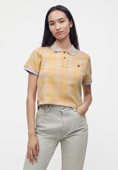 Jeune femme portant un polo court à carreaux jaunes avec des bordures bleues et un jean clair taille haute, posant la main dans la poche.