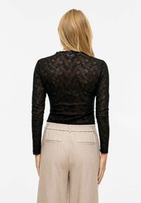 Czarna bluzka z długim rękawem z koronki z delikatnym kwiatowym wzorem, zestawiona z jasnobeżowymi plisowanymi spodniami z elastycznym paskiem w talii i kieszeniami.