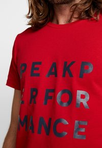 Camiseta de algodón roja con un atrevido texto impreso en negro que dice "PEAK PERFORMANCE". Cuello redondo, mangas cortas, textura suave.