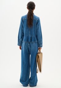 Denim jumpsuit met een peplum taille, lange mouwen en wijde pijpen. Geaccentueerd met een beige tote bag. Achteraanzicht tegen een witte achtergrond.