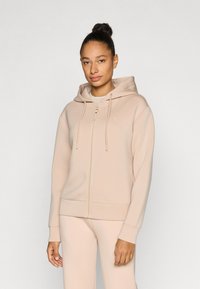 Φωτεινή μπεζ ζακέτα με φερμουάρ, κατασκευασμένη από απαλό ύφασμα. Διαθέτει κουκούλα, κορδόνι και μπροστινές τσέπες. Ομαλή υφή με ελάχιστη λεπτομέρεια εταιρικού σήματος.