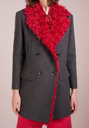 Manteau gris foncé à double boutonnage avec un grand col en fausse fourrure rouge et doublure, porté avec un pantalon rouge assorti.