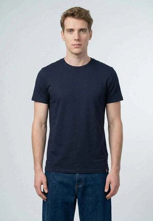 Junger Mann mit kurzen lockigen Haaren, der ein schlichtes dunkelblaues T-Shirt und blaue Jeans trägt, vor einem hellgrauen Hintergrund stehend.