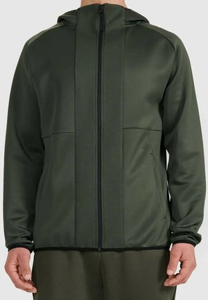 Man draagt een olijfgroene hoodie met rits en een gestructureerd paneel aan de voorkant en bijpassende broek, staand met ontspannen handen.