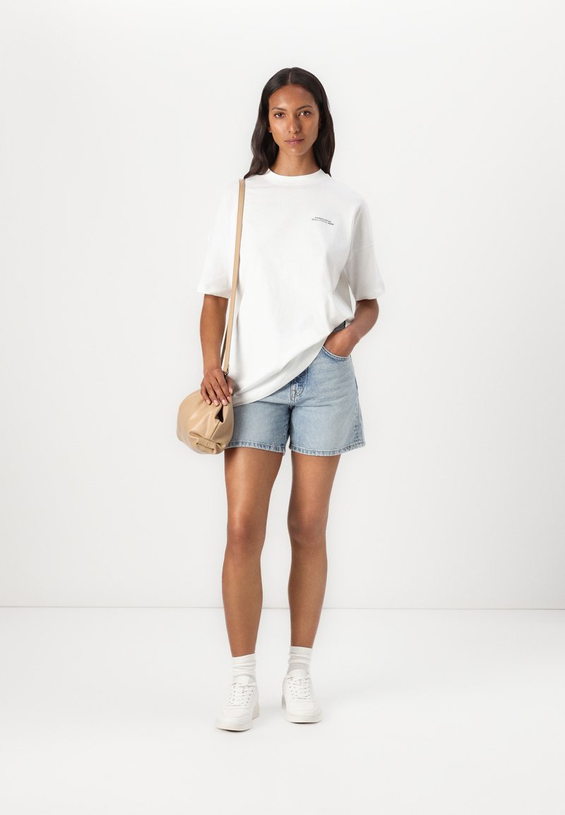 Weißes Baumwoll-T-Shirt mit kurzen Ärmeln und kleiner Grafik, kombiniert mit hellblauen Denim-Shorts. Dazu kommt eine beigefarbene Umhängetasche und weiße Sneakers.
