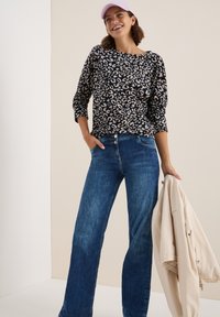 Haut à manches longues en imprimé léopard noir et beige associé à un jean bleu. Veste de couleur claire tenue d'une main. Look décontracté et ample.