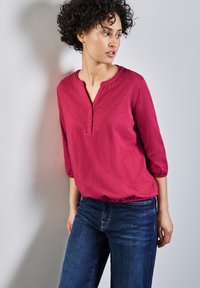 Fuchsiová blůza s tříčtvrtečními rukávy, henley výstřihem a knoflíkovou légou; nošena s modrými džíny; hladká textilní struktura.