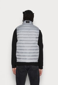 Graue, gepolsterte Weste mit horizontalen Nähten, hohem Kragen und Calvin Klein-Label auf der Rückseite, getragen über einem schwarzen Langarmshirt.