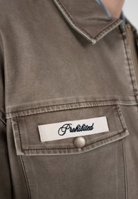 Brun jakkelomme med metal trykknap og hvidt stofmærke broderet med ordet "Prohibited" i sort kursiv skrift.