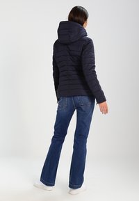 Kobieta stoi tyłem, ubrana w czarną pikowaną kurtkę z kapturem, niebieskie dżinsy i białe trampki na jednolitym tle.