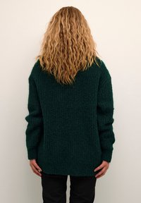 Persoon met lang krullend blond haar, gekleed in een donkergroene gebreide trui en zwarte broek, staande met het gezicht naar een effen lichte muur.
