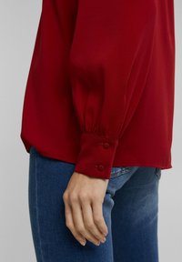 Rød, langærmet bluse med en glat overflade, der har knappede manchetter og en afslappet pasform, som er parret med blå jeans.