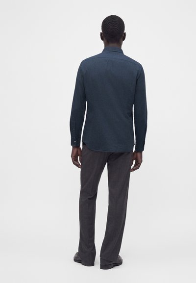 Homme aux cheveux courts portant une chemise bleu foncé à manches longues et un pantalon gris foncé, debout dos à un fond uni.