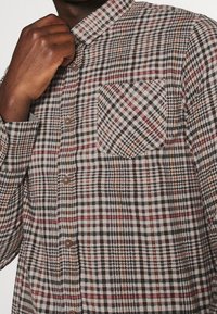 Persona de piel oscura que lleva una camisa de botón de manga larga con un patrón a cuadros en rojo, negro y beige, sosteniendo el cuello con una mano.