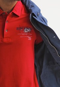 Homme portant un polo rouge avec un petit motif imprimé et une veste bleu marine partiellement posée sur l'épaule, sur fond blanc.