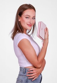 Light pink wallet with zipper closure, smooth texture, rounded edges, and a decorative accent. Held by a person in a fitted white top.