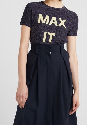Persoon draagt een marineblauw glinsterend T-shirt met de tekst "MAX IT" en een hoog geplooide donkerblauwe rok tegen een effen achtergrond.