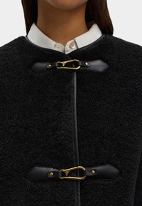 Cappotto esterno in lana nera e testurizzata con colletto alto, dotato di chiusure in metallo dorato e accenti in pelle nera. Camicia bianca button-down sotto.