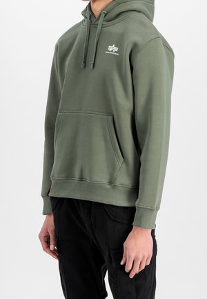 Uomo che indossa una felpa con cappuccio Alpha Industries verde oliva con tasca frontale e pantaloni cargo neri su sfondo semplice.