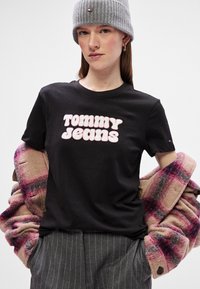 Μαύρο T-shirt με το "TOMMY JEANS" σε ροζ φουσκωτά γράμματα. Ένα μοντέλο φοράει ένα γκρι πλεκτό σκουφί και ένα oversized μπουφάν σε καρό εκρού και ροζ.