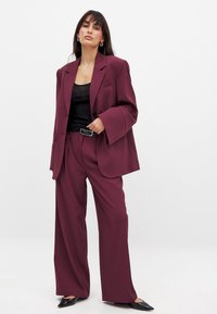 Burgundska tailoring blazer in široke hlače, v kombinaciji s črno majico brez rokavov. Blazer ima strukturirane ramena in žepne preklopne žepke.