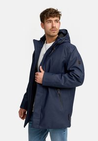 Marineblauwe waterdichte jas met een capuchon, ritszakken, trekkoordaccenten en een gladde textuur. Model draagt het over een licht shirt.