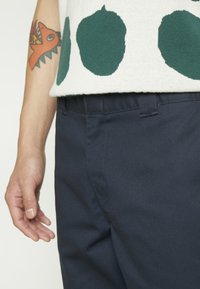 Pantaloni din bumbac bleumarin cu o textură netedă, având brâuri și buzunare laterale. Partea de sus afișează un pulover crem cu modele circulare verzi.