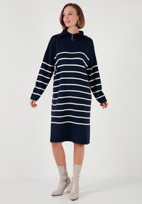 Rochie tricotată bleumarin cu dungi orizontale albe, guler cu fermoar și mâneci lungi. Modelul poartă botine lucioase bej cu toc accentuat.