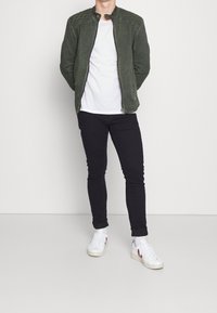 Zelená semišová bomber bunda s prošívanými akcenty na ramenou, kombinovaná s bílým tričkem, černými úzkými džíny a bílými teniskami.