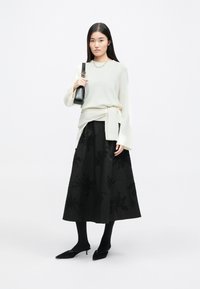 Sweater cinza claro com mangas largas e cintura atada, combinado com uma saia longa preta adornada com padrões florais bordados e lantejoulas.