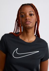 Jovem mulher com longas tranças vermelhas, vestindo uma t-shirt preta com um grande logótipo da Nike em branco sobre um fundo branco.
