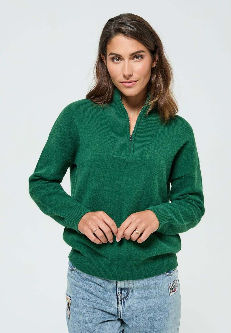 Pull en tricot vert avec un col zippé à mi-hauteur, texture côtelée et manches retroussées. Associé à un jean bleu clair avec des patches.