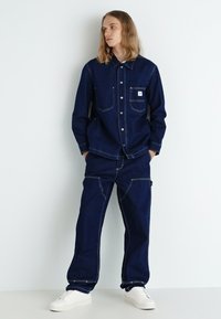 Completo in denim blu scuro, composto da camicia e pantaloni, con chiusura a bottoni, tasca frontale e cuciture bianche a contrasto. Abbinato a sneakers bianche.