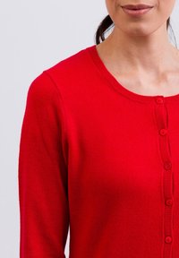 Červený pletený kardigan s kulatým výstřihem, zapínáním na knoflíky a dlouhými rukávy. Hladká textura s plochým pletením a žebrovanými detaily.