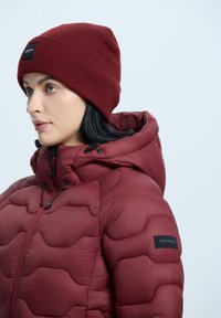 Bordowa kurtka puchowa o pikowanej fakturze i kapturze, zestawiona z dopasowaną czapką z ribbowej dzianiny. Posiada naszywkę z logo na rękawie kurtki.
